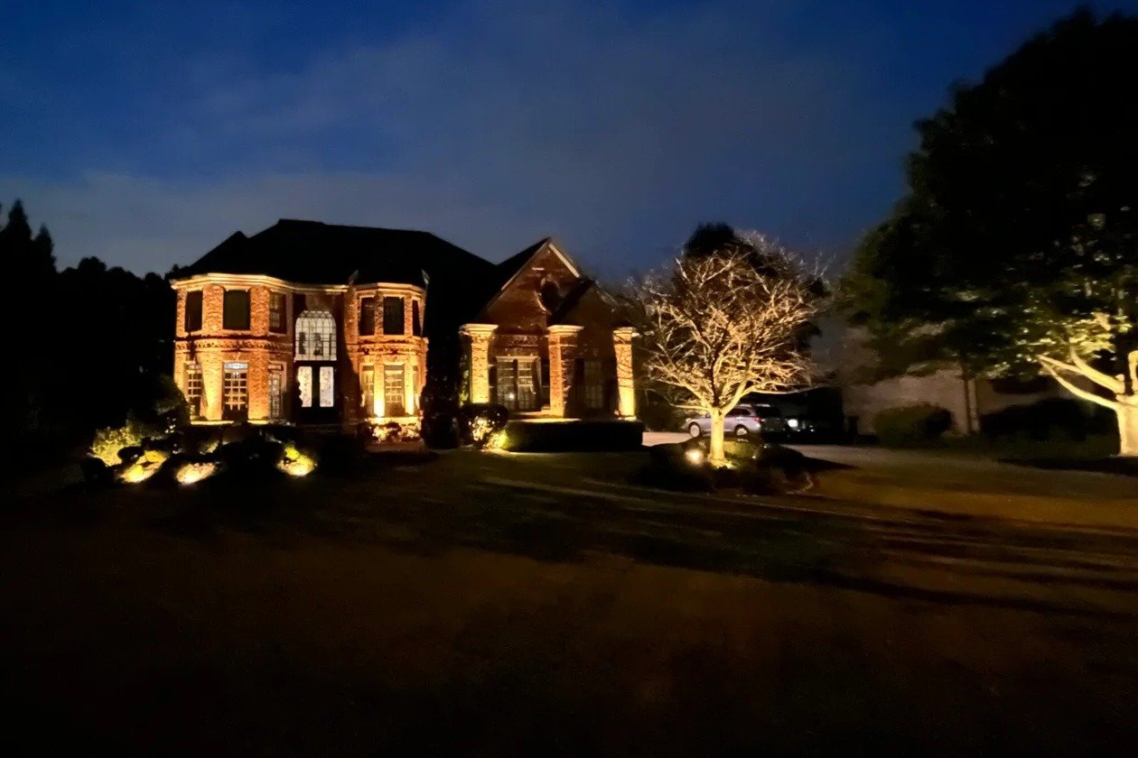 Custom brick home at dusk with façade uplighting, an uplit specimen tree, and landscape accent lighting